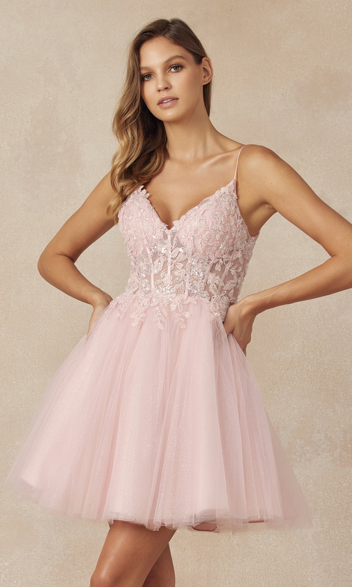 Wearing the Sheer-Bodice Short A-Line Homecoming Dress 863, a woman stands confidently with hands on hips in a light pink, corset-style v-neck dress featuring a fitted lace bodice and flared tulle skirt against a beige background.